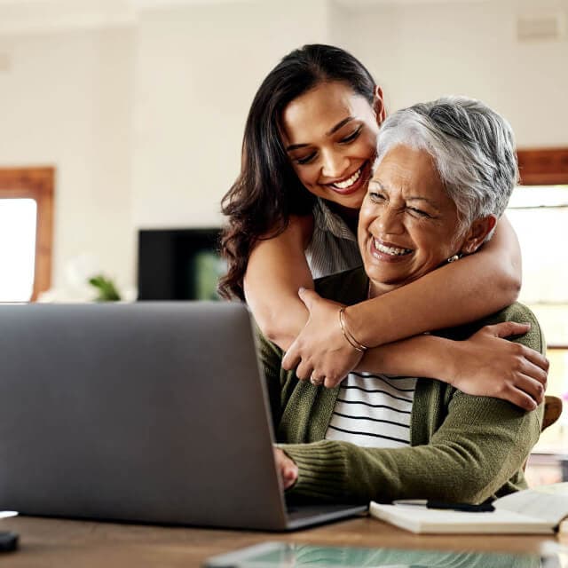 Jovem sorrindo abraçando sua mãe em frente ao computador